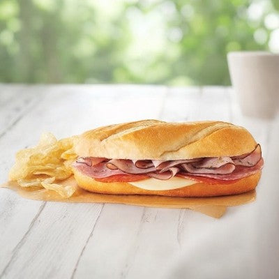 A delicious Italian Style Sub from E.A. Sween, featuring premium Italian cold cuts and cheese on a freshly baked roll, is served with potato chips on a wooden table. A blurred green background and white cup enhance the inviting scene. Ships frozen with 30 days shelf life in the cooler!.