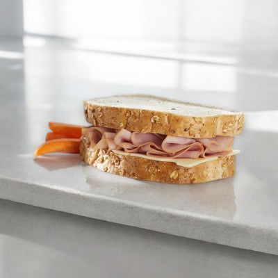 The E.A. Sween Ham & Swiss Sandwich features premium ham on rustic oat-topped wheat bread, complemented by three carrot sticks, elegantly displayed on a marble surface.