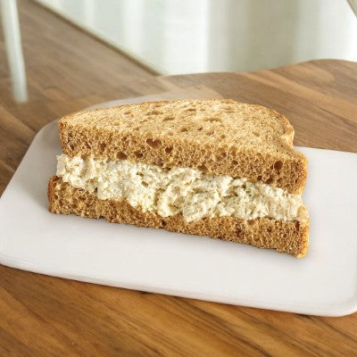 The E.A. Sween Ranch Chicken Salad Sandwich on multigrain bread sits elegantly on a white plate atop a rustic wood table. Ships frozen, lasts 30 days in the cooler!.