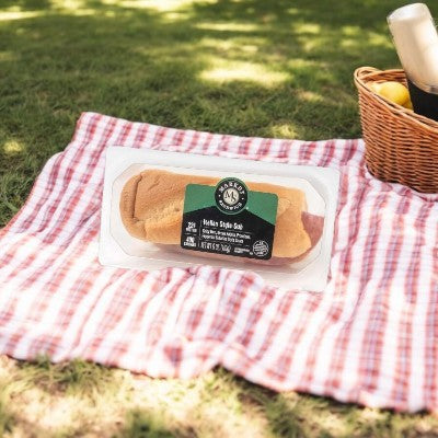 An Italian Style Sub from E.A. Sween, freshly baked and featuring premium Italian cold cuts, rests on a checkered picnic blanket. A nearby wicker basket full of apples completes the inviting outdoor feast setup.