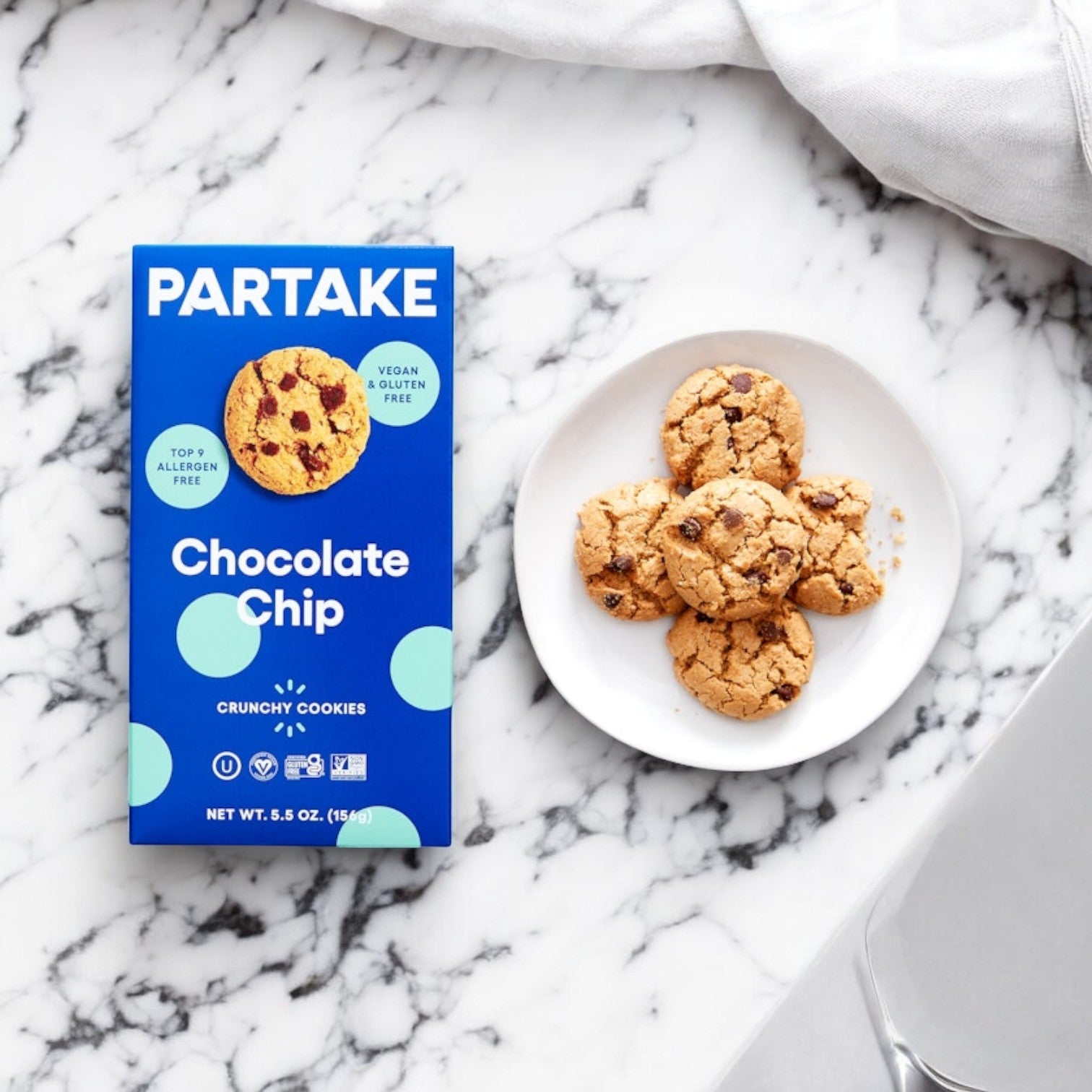 A box of Partake Gluten Free Vegan Chocolate Chip Cookies, 5.5 oz - 1 Count, made with inclusivity in mind, rests beside a white plate holding five cookies on a marble surface.