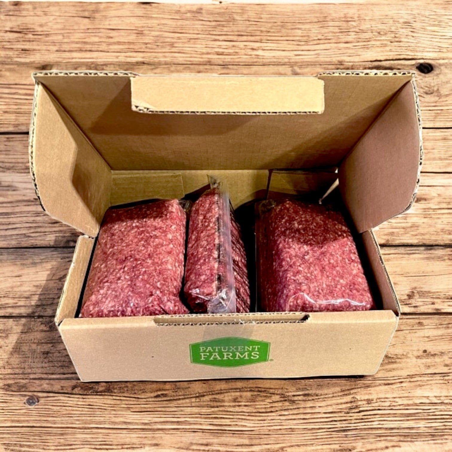 An open box of Patuxent Farms Halal Ground Lamb 80/20 (raw, frozen, vacuum-packed), with multiple packs totaling 10 lbs, is displayed on a wooden surface.