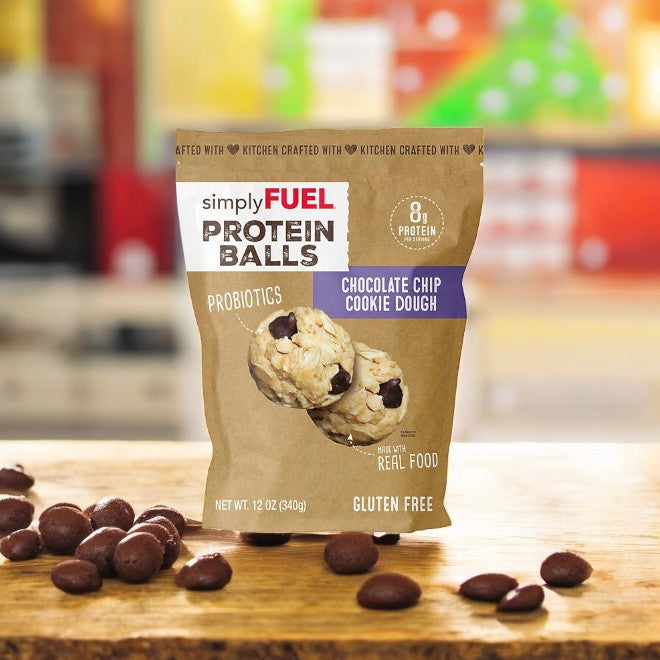 A 12 oz bag of Simple Fuel Chocolate Chip Cookie Dough Protein Balls, featuring plant-based protein, probiotics, and gluten-free ingredients, sits on a wooden table surrounded by scattered chocolate balls.