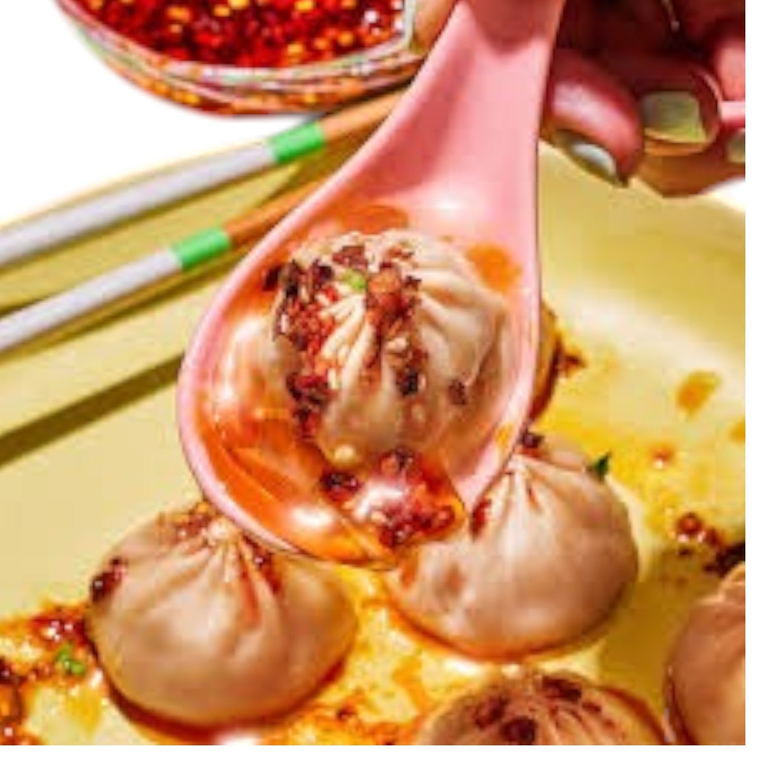 A pink spoon holds a Mila 3 Pack Soup Dumpling Combo dumpling with chili oil above a dumpling plate, accompanied by chopsticks and a bowl of flavorful chili oil in the background.