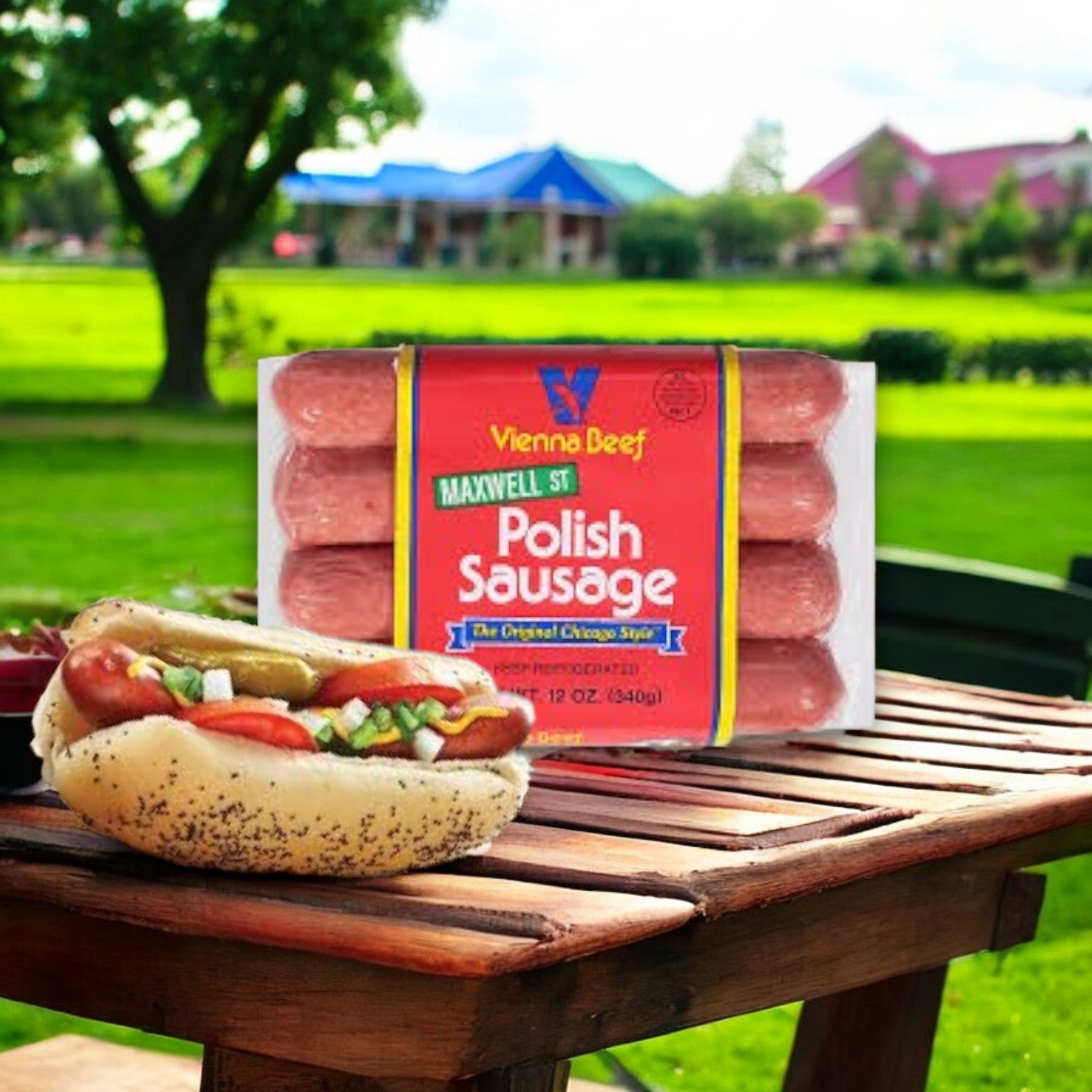 A Vienna Beef Polish Sausage-12 oz-1 Count sits on the picnic table, while another in a seeded bun, topped with condiments, takes center stage. In the background, a vibrant park and colorful buildings are visible.