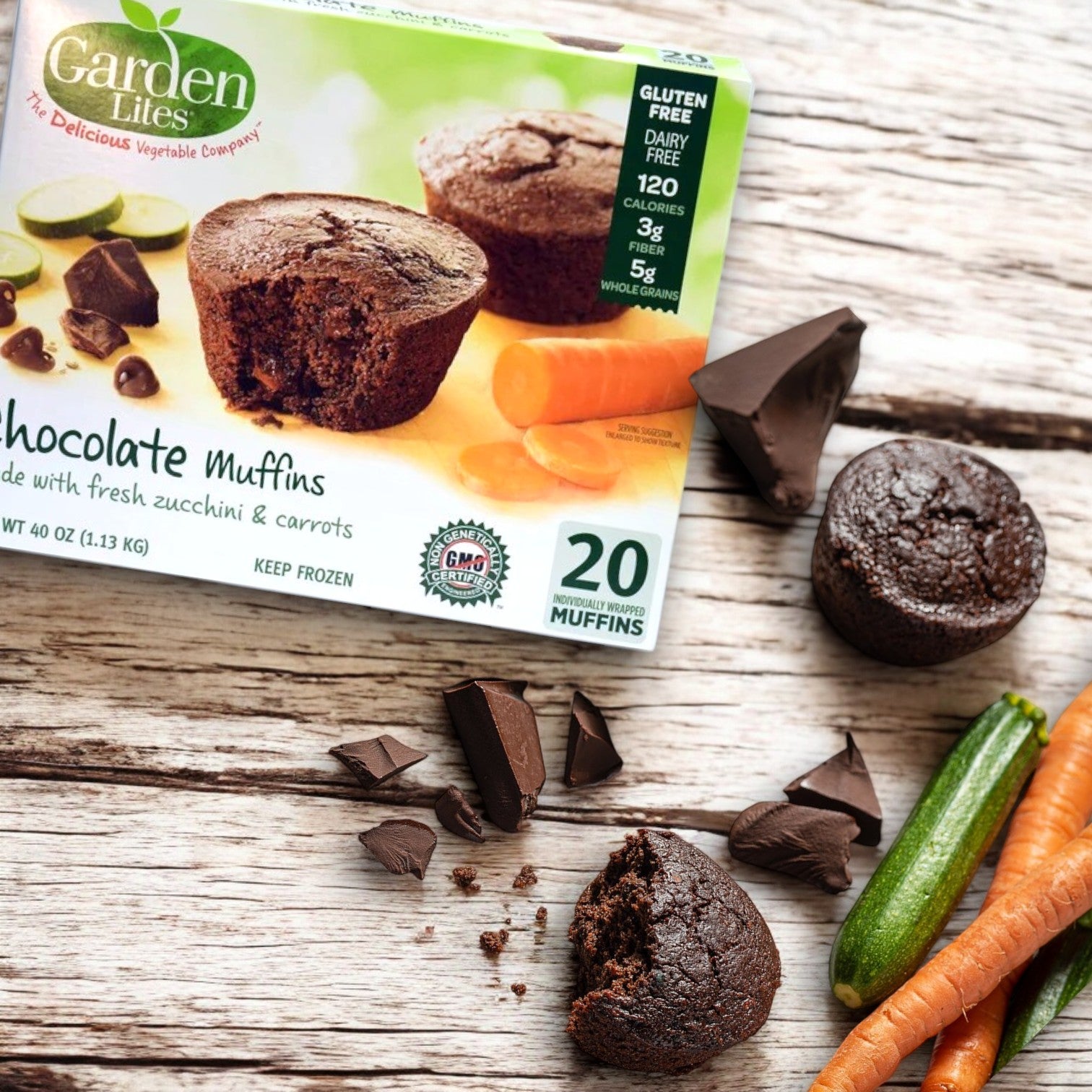 A 40 oz. box of Veggies Made Great Garden Lites Chocolate Muffins sits on a wooden surface, surrounded by rich chocolate pieces, whole zucchini, and carrots. These deliciously moist, veggie-packed treats are gluten-free indulgences without the guilt.