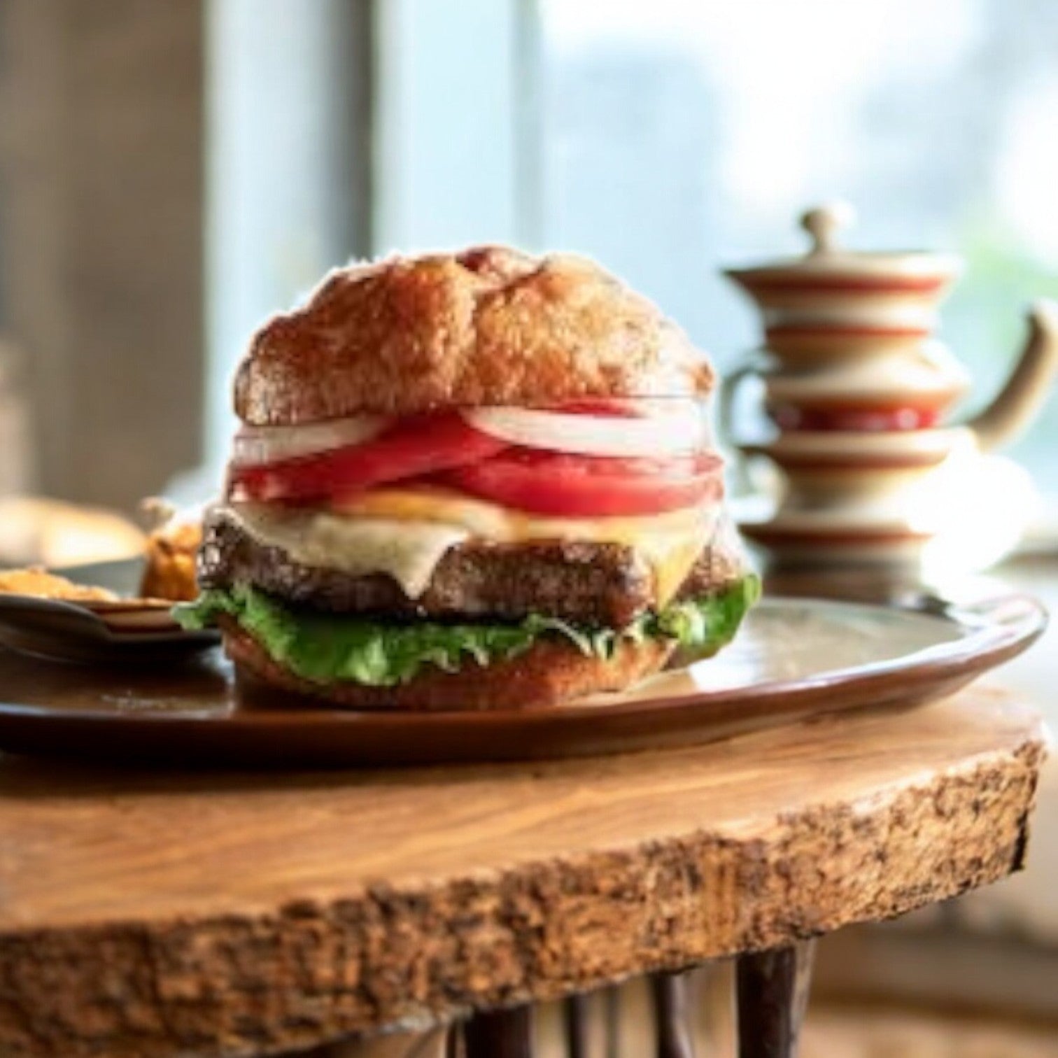 A Bubba Burger Original from a 6-count frozen, 32 oz pack sits enticingly topped with lettuce, tomato, onion, and cheese on a wooden plate next to a small dish, as a teapot and cups blur softly in the background.