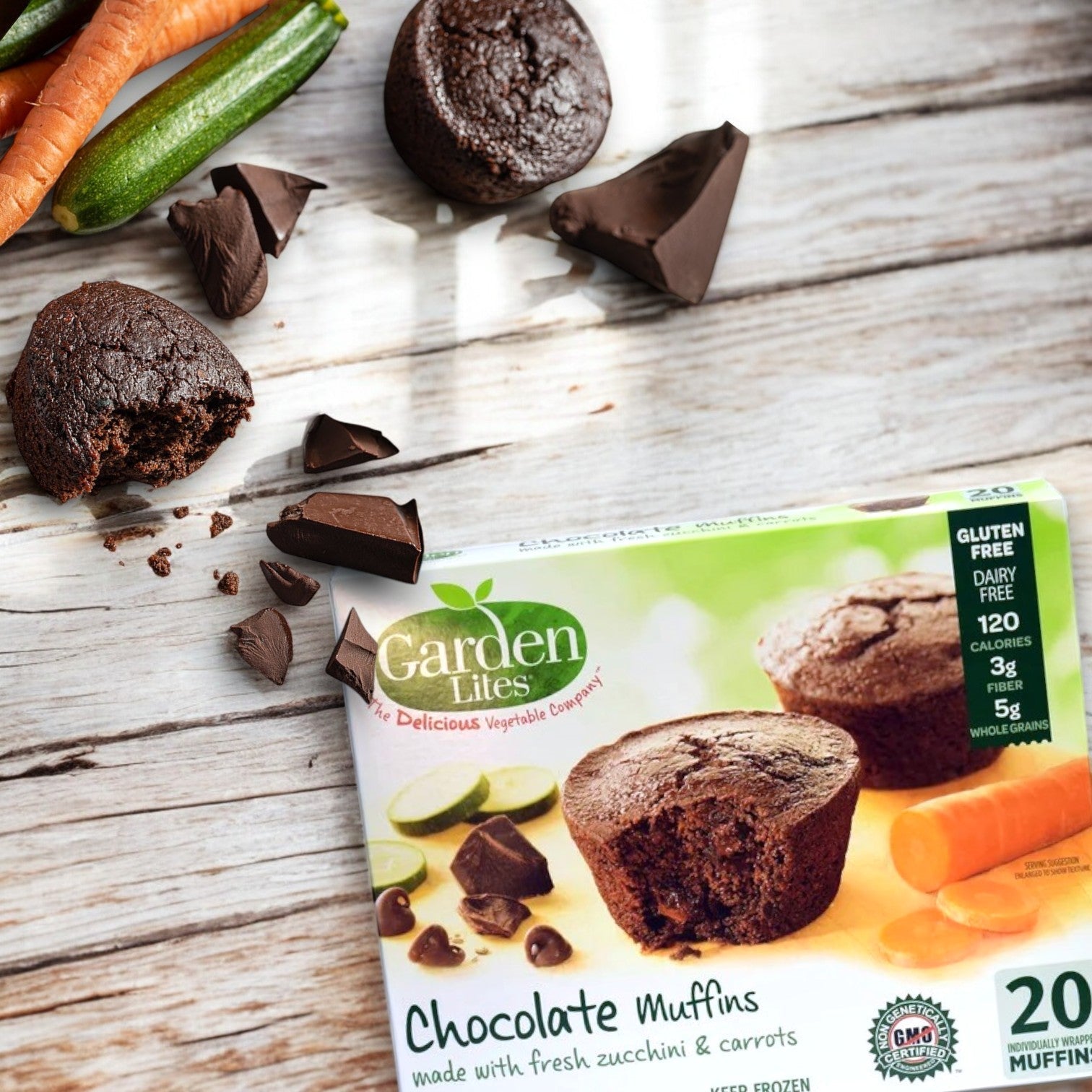 A box of Veggies Made Great's Garden Lites Chocolate Muffins – Deliciously Indulgent & Veggie-Packed (120 oz.) rests on a wooden surface, surrounded by muffins, chocolate pieces, zucchini, and carrots. The packaging proudly showcases its dairy-free and gluten-free contents.