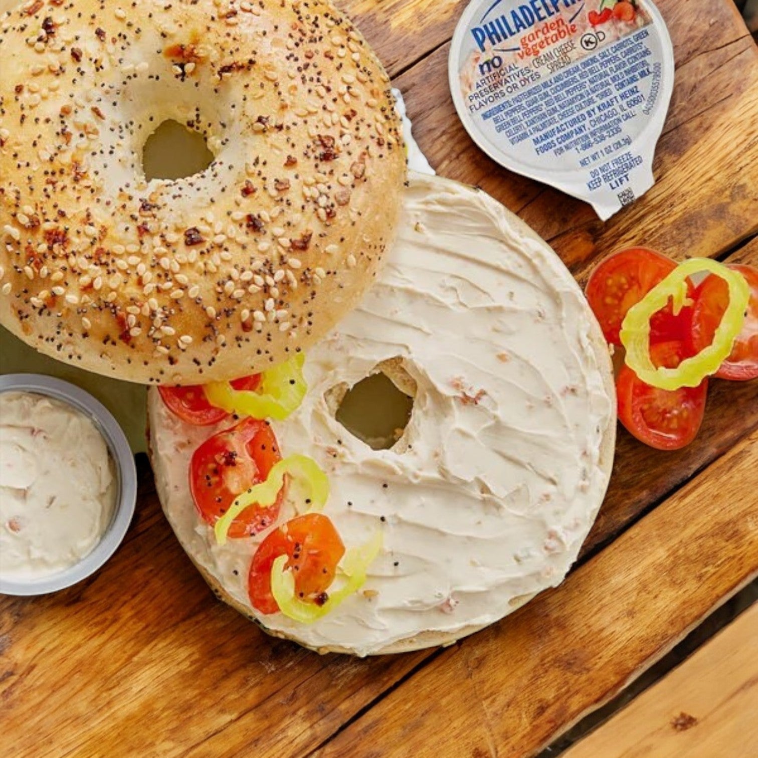 A wooden surface holds a half bagel with Philadelphia Garden Vegetable Cream Cheese, tomato slices, and pepper rings. The other half features seeds. Nearby are tomatoes and a Kraft Philadelphia Garden Vegetable Cream Cheese container from the 100 Pack of 1 oz. servings.