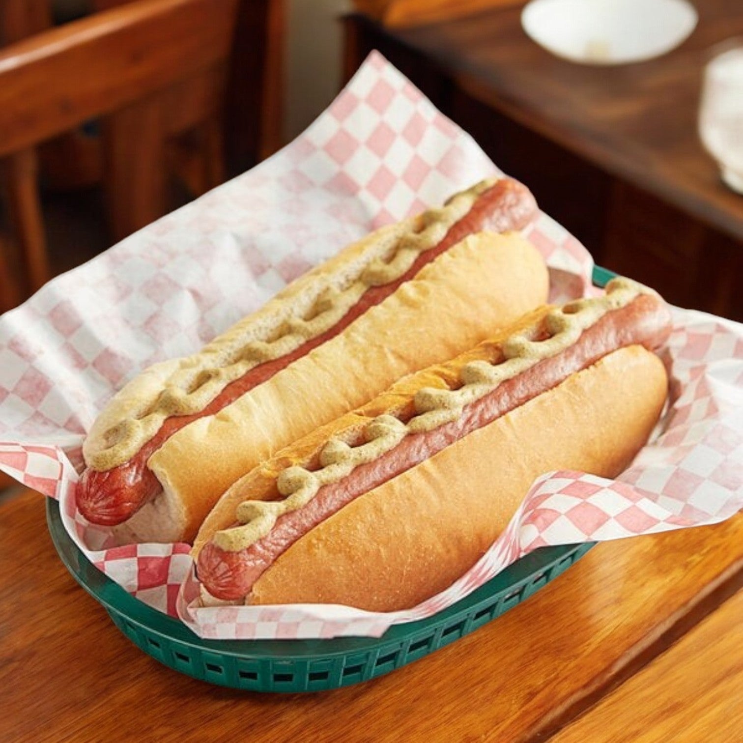 Two 8" bun length hot dogs with mustard in buns sit on checkered paper in a green basket on a wooden table. Made with Nathan's Famous 8" Bun Length Beef Franks, these premium all-beef hot dogs are perfect for any occasion.