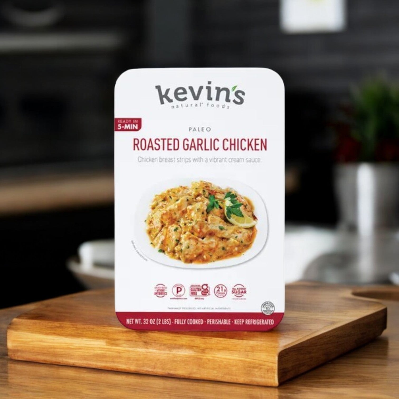 Kevin's 32 oz. Roasted Garlic Chicken packaging features a savory dish image with text highlights like gluten-free, paleo-friendly, ready in 5 mins, and fully cooked, all elegantly displayed on a rustic wooden board.