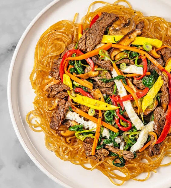 A dish of japchae with sweet potato noodles, FoodHaul marinated Korean Flank Steak, red peppers, mushrooms, spinach, and strips of egg is elegantly presented on a white plate—a true delight from Korean cuisine.