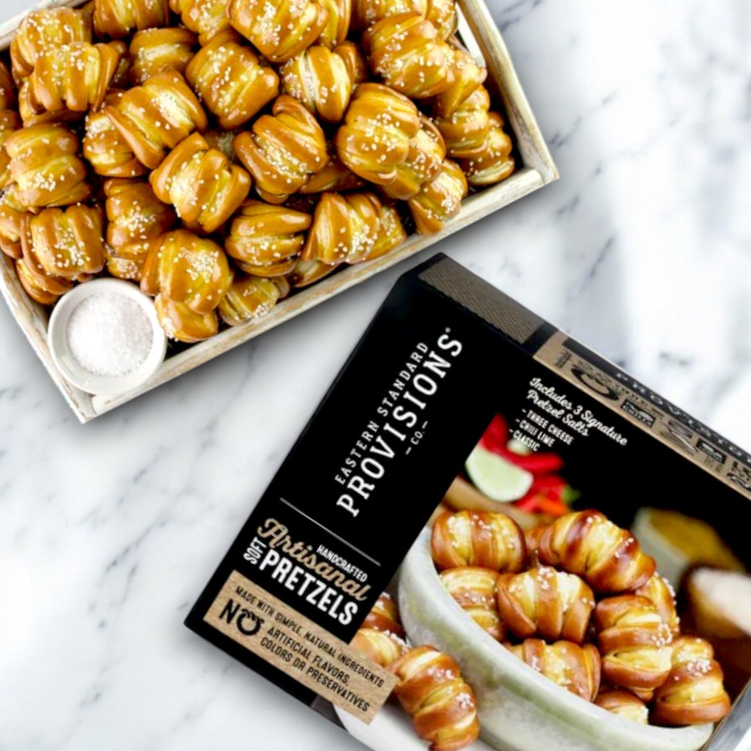 A tray of Eastern Standard Provisions Artisanal Soft Pretzel Bites, crafted with care and topped with coarse salt, sits beside a bowl of salt. Below is a box labeled "Eastern Standard Provisions," ideal for versatile snacking.