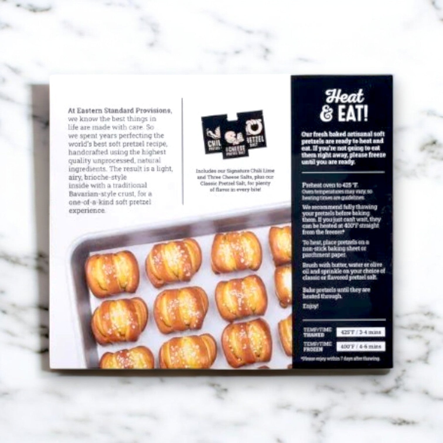 A box of Eastern Standard Provisions Soft Artisanal Handcrafted Pretzels, weighing 13.1 oz., is displayed on marble, highlighting the artisanal craftsmanship. The packaging features heating instructions and mentions gourmet dipping salts for versatile snacking.