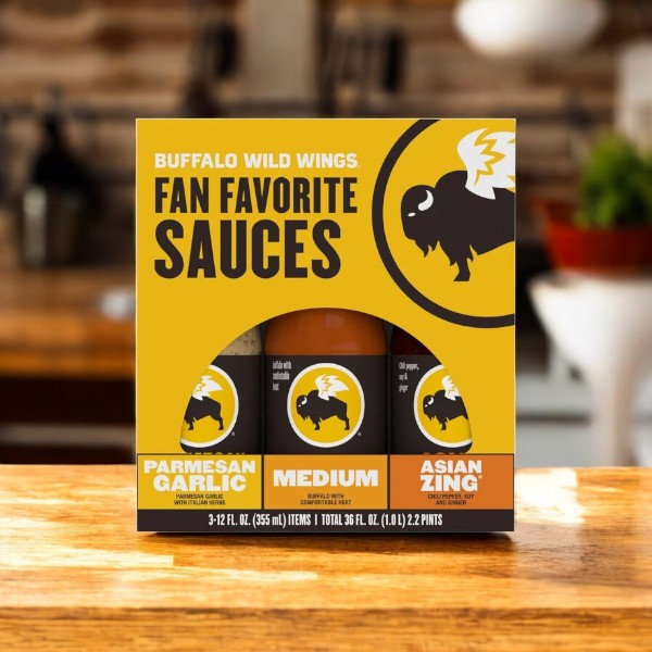 Explore the Buffalo Wild Wings Fan Favorite Variety Pack with Parmesan Garlic, Medium, and Asian Zing sauces on a wooden table. A blurred kitchen backdrop with jars and a plant highlights these vibrant and tantalizing flavors.