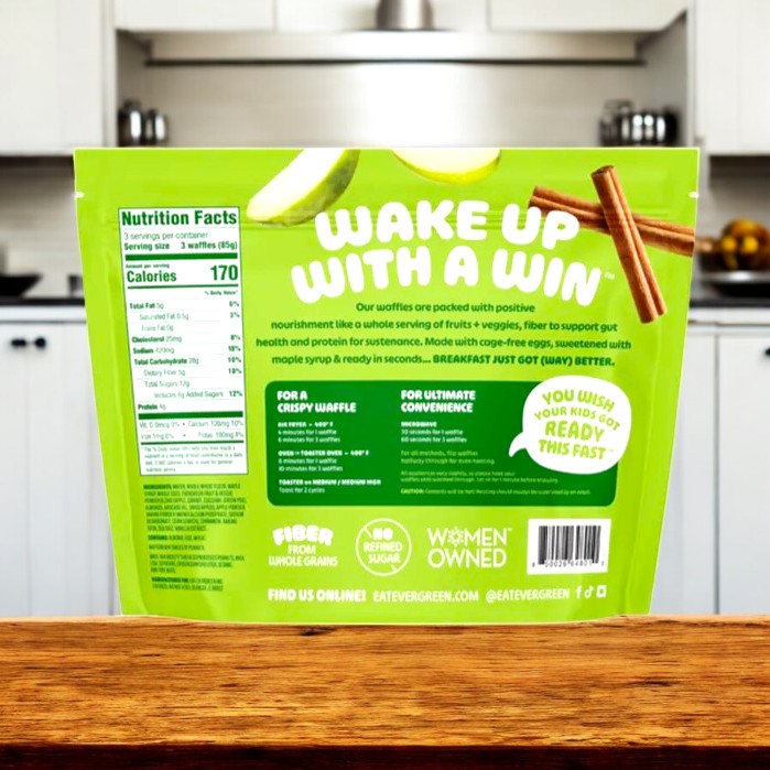 A green Evergreen package of Apple Cinnamon Cobbler Frozen Mini Waffles (9oz, 3 count) stands on a wooden counter in a kitchen, displaying nutrition facts and the slogan "Wake Up With A Win"—ideal for a sweet snack anytime.