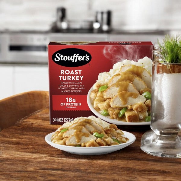 A Stouffer's Roast Turkey meal, 9.625 oz, featuring homestyle gravy, stuffing, and mashed potatoes, is placed beside a served plate on a wooden table in a cozy kitchen setting.