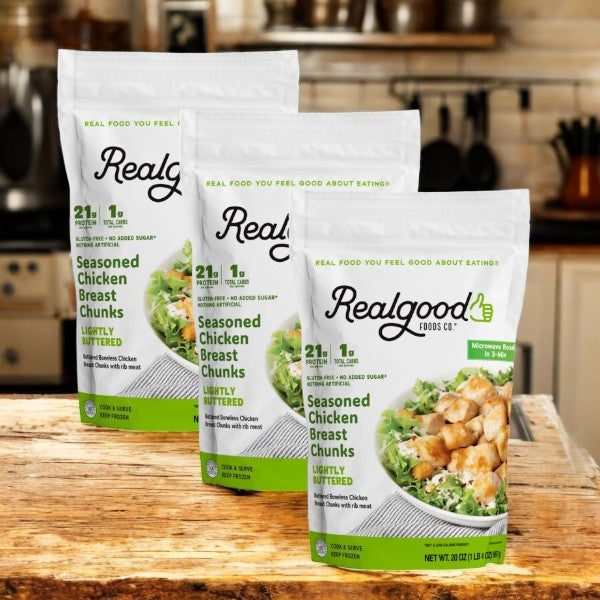 Three bags of Real Good Foods Lightly Buttered Chicken Breast Chunks sit invitingly on a wooden table in a cozy kitchen. Each 20 oz bag features images of savory chicken chunks and crisp salad, emphasizing their keto-friendly appeal.