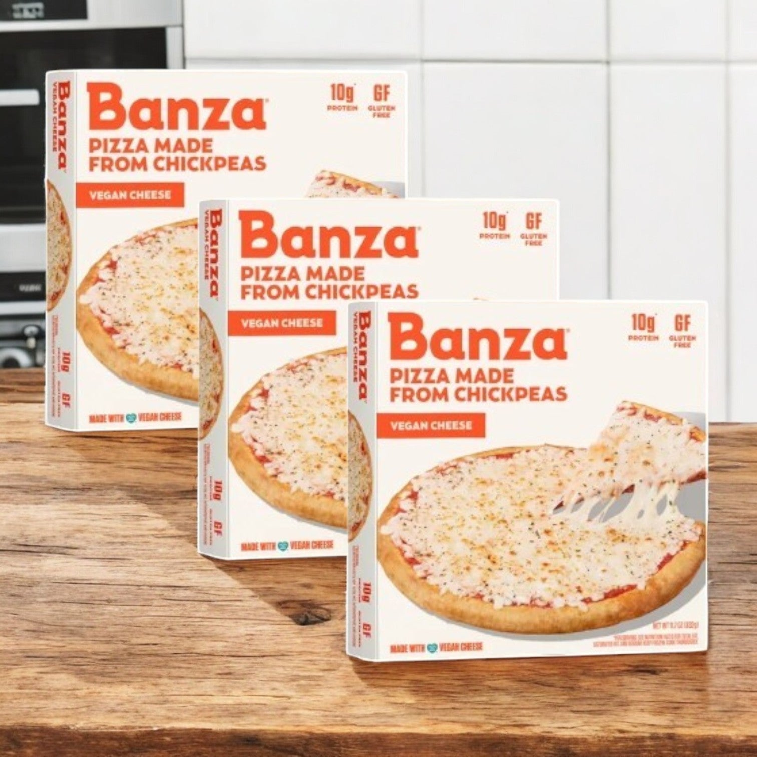 Three boxes of Banza Vegan Cheese Pizza, with a chickpea-based gluten-free crust, arranged in a staggered formation on a wooden countertop against a kitchen backsplash.