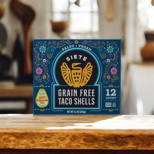 A box of Siete Taco Shells, Grain Free-12 Count-1 Pack, rests on a wooden table. Adorned with colorful floral patterns, they're gluten-free and paleo-friendly. Made with 100% avocado oil, these shells are a tasty and healthy choice for your next meal.