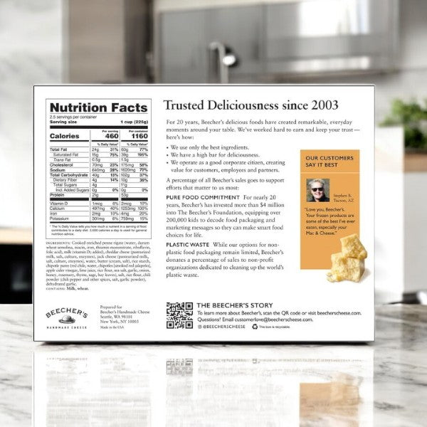The back of Beecher's Handmade Cheese "World's Best" Mac & Cheese box features nutrition facts, a glowing customer testimonial, and company info in a cozy kitchen setting. Discover why aged cheddar is used to craft this beloved dish by Beecher's.