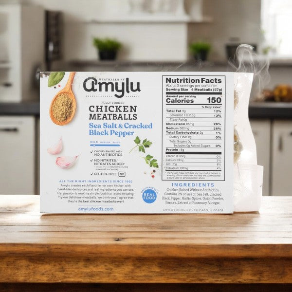 On a wooden kitchen counter sits a 10 oz box of Amylu Chicken Meatballs, fully cooked and seasoned with sea salt and cracked black pepper. The nutrition facts and high-quality chicken ingredients are visible against a cozy, blurred kitchen background.