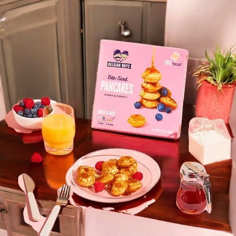 A breakfast setup features Belgian Boys Refrigerated Bite-Sized Pancakes on a plate, drizzled with syrup, alongside a glass of orange juice and fresh berries. A box labeled "Belgian Boys Bite-Sized Pancakes-1 Count" is displayed in the background.