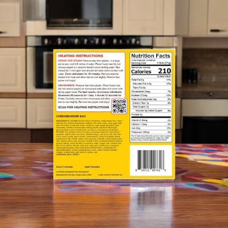 A Wow Bao Cheeseburger Bao-1 Count box sits on a wooden table against a kitchen backdrop, highlighting its fusion of bold flavors. The packaging features heating instructions, a QR code, nutrition facts, and a product description.