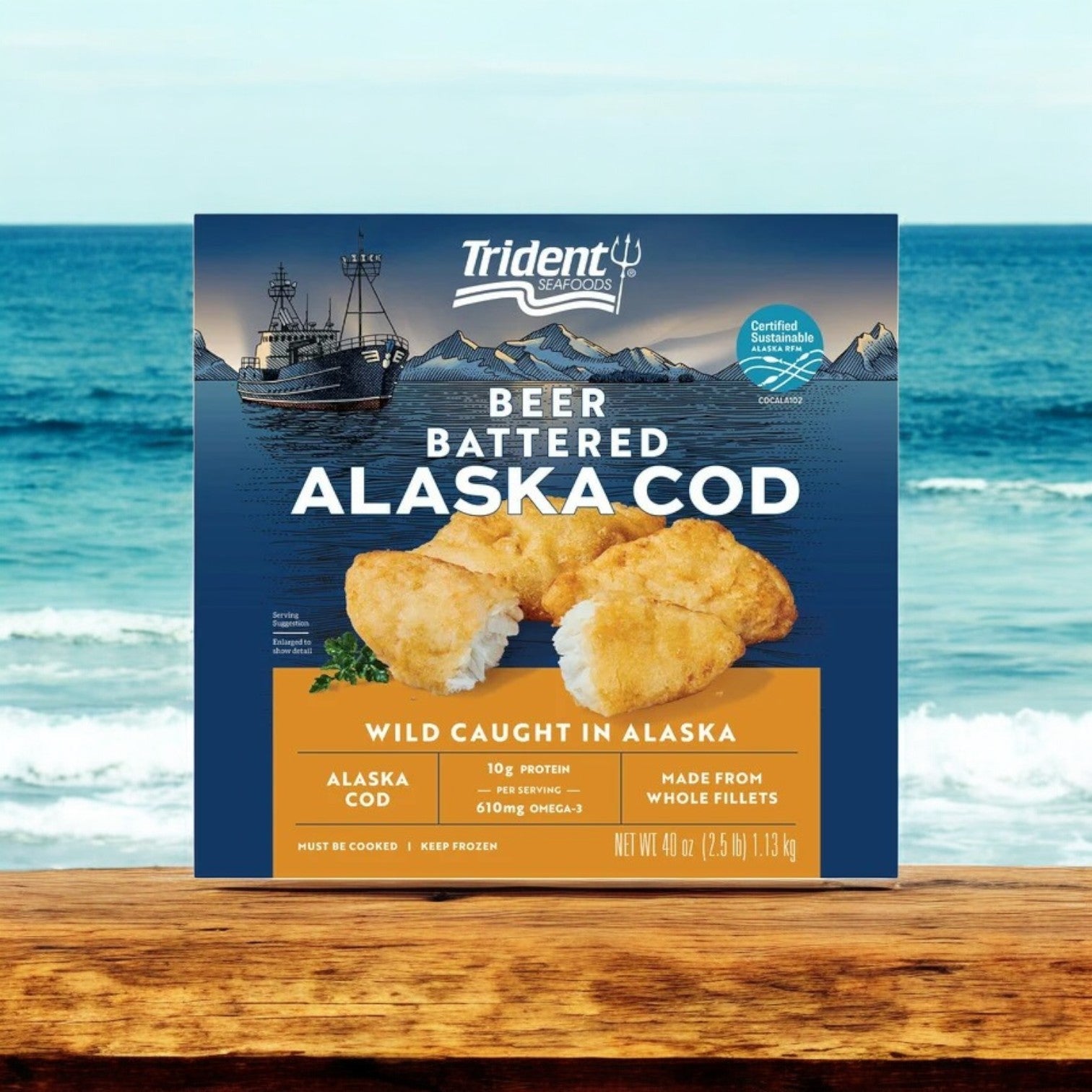 Trident Seafoods Wild Alaskan Beer Battered Cod (3 boxes, 7.5 lbs) on a wooden table with the ocean and a fishing boat in the background.