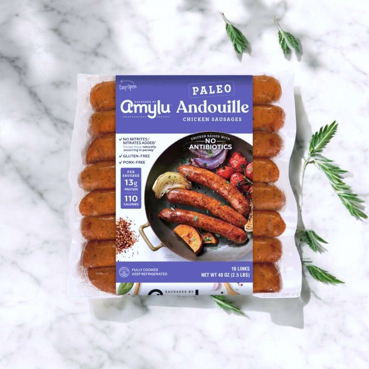 A pack of Amylu Paleo Andouille Sausages is displayed on a white surface with greenery. These sausages, featuring authentic flavor, are labeled nitrate-free, gluten-free, and paleo-friendly. Available as a 16 count in 3 packs by Amylu.