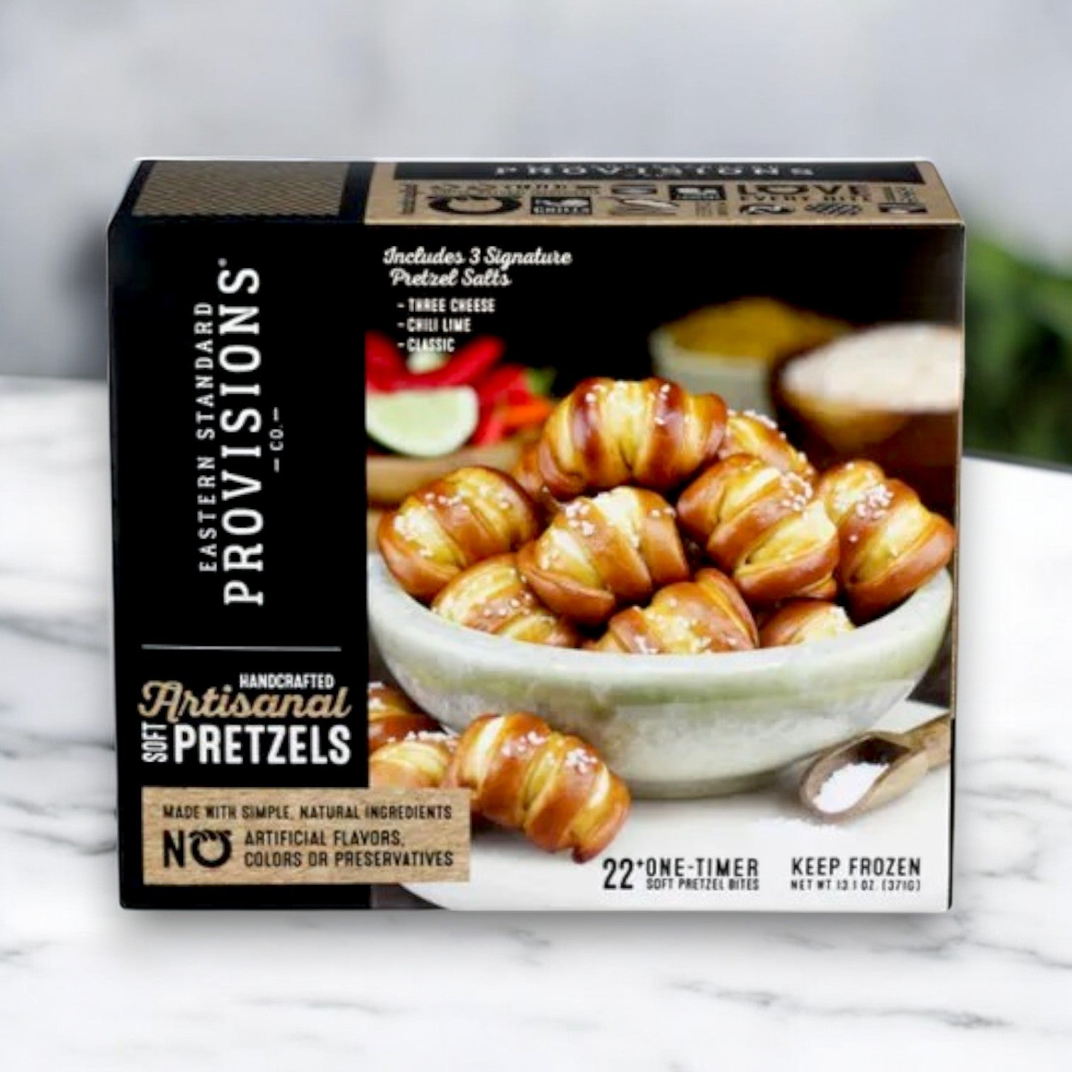 A 13.1 oz. box of Eastern Standard Provisions Pretzels sits on a marble surface, exemplifying versatile snacking with its natural ingredients and preservative-free promise, while pretzels are showcased elegantly in a white dish.