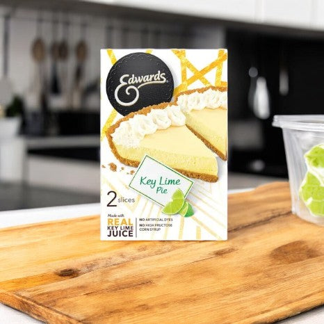 An Edwards Key Lime Pie-1 Count from Edward's Desserts, with its signature graham cracker crust, is elegantly displayed on a wooden board, kitchen utensils in the background.