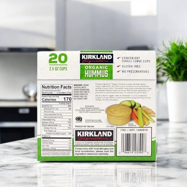 A box of 20 Kirkland Signature Organic Hummus single-serve cups sits on the kitchen counter, labeled gluten-free and made with organic chickpeas, containing no preservatives.