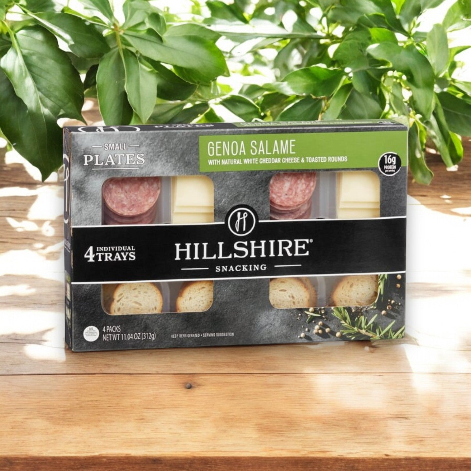 A box of Hill Shire Snacking Plates, White Cheddar and Genoa Salame (4 ct) is elegantly displayed on a wooden surface with green leaves in the background.