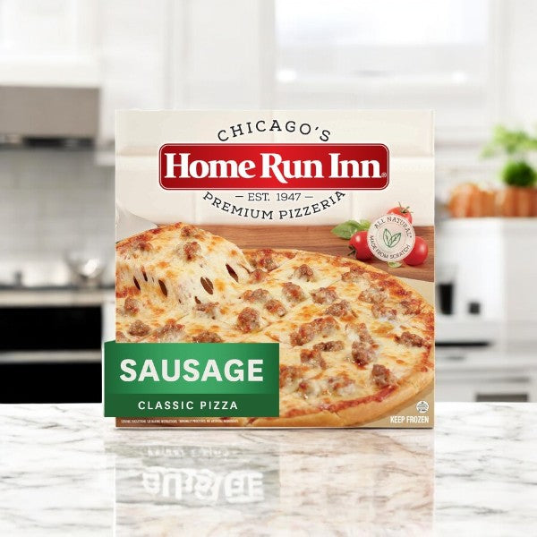 A box of Home Run Inn Family Sausage Pizza (30 oz., 1 count) is displayed on a marble kitchen countertop. The packaging features an image of the pizza, emphasizing its mozzarella cheese, buttery flaky crust, and brand details.