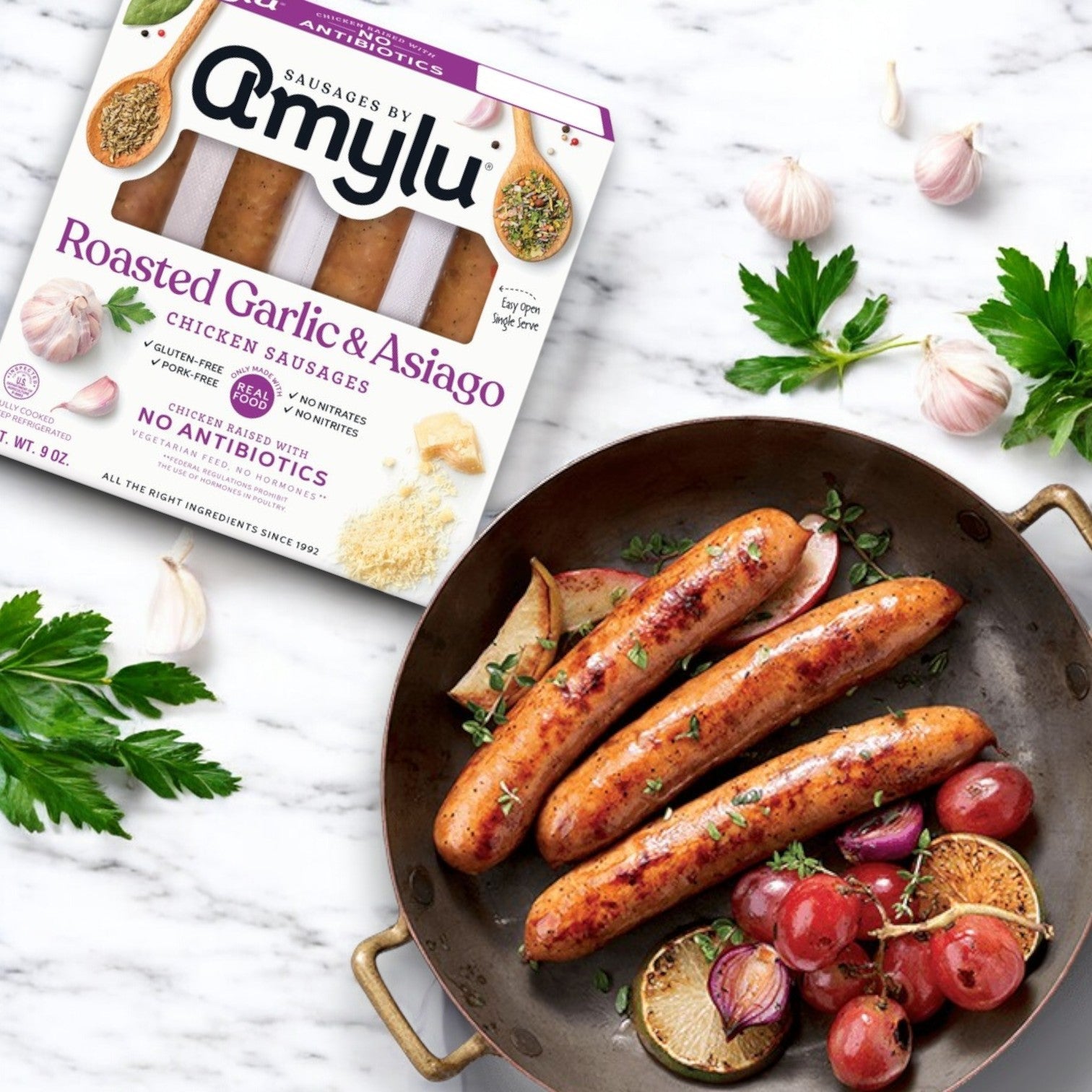 Amylu Roasted Garlic & Asiago Chicken Sausage - 3 Packs, displayed beside a pan of cooked sausages, garnished with herbs, lemon slices, and roasted grapes. Garlic cloves and parsley are scattered around.