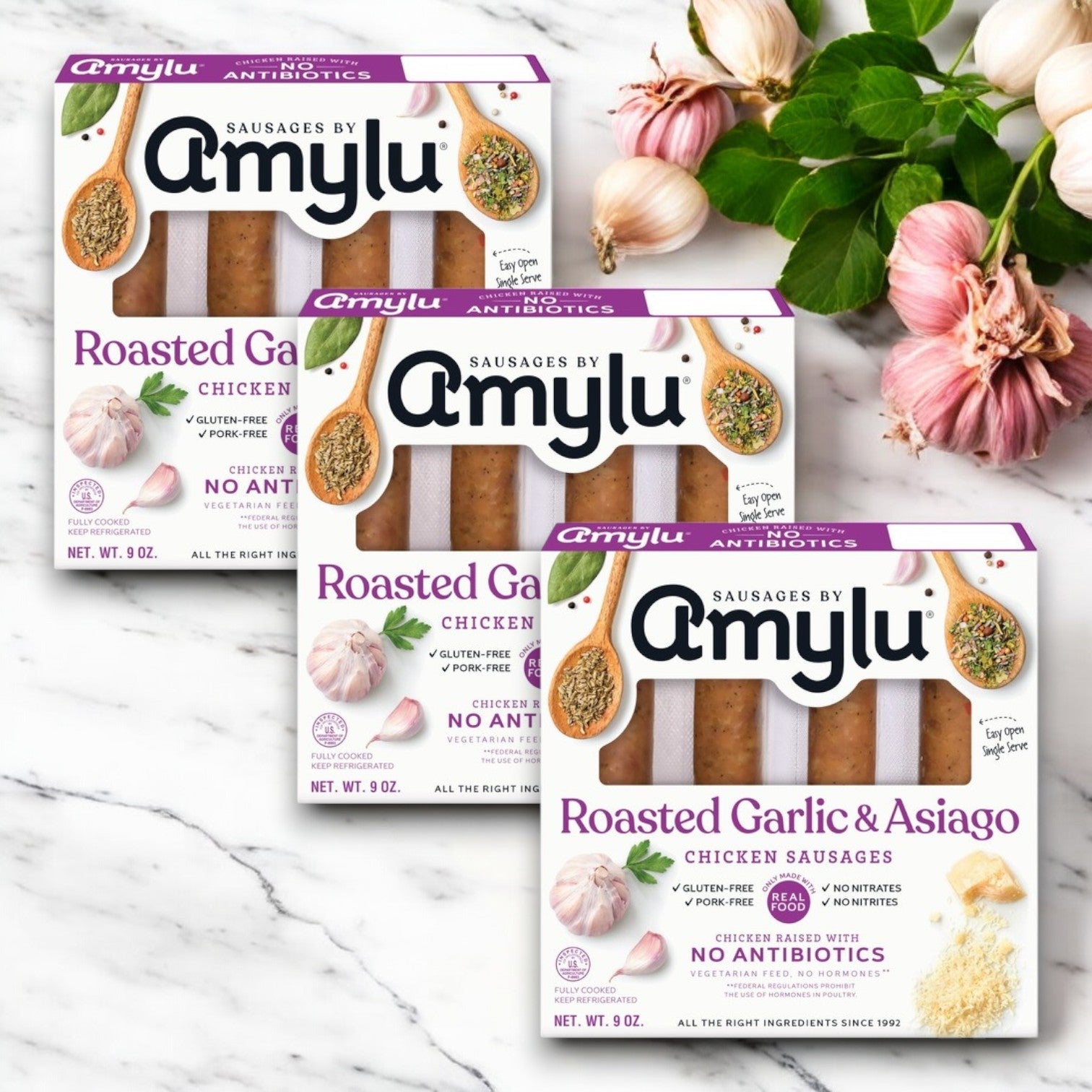 Amylu Roasted Garlic & Asiago Chicken Sausage - 3 Packs rest on a marble surface, accented by garlic cloves and herbs, showcasing the savory blend of chicken.