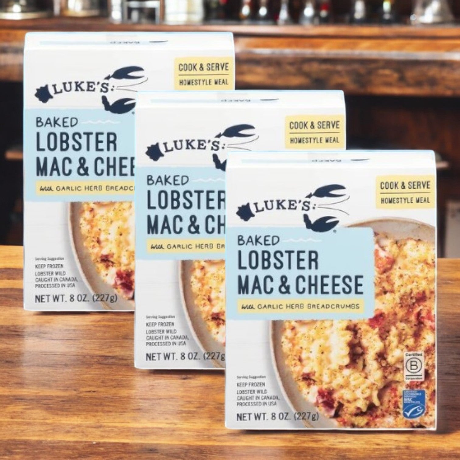 Three boxes of Luke's Organic Baked Lobster Mac & Cheese- 3 Count are elegantly displayed on a wooden counter, highlighting this gourmet dish topped with garlic herb breadcrumbs.