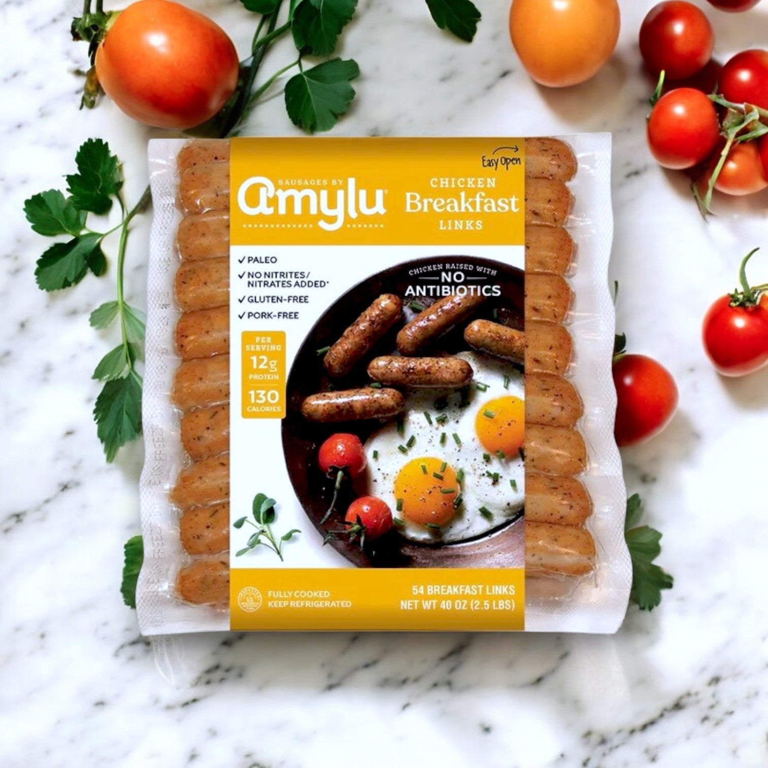 A 2.5 lbs pack of Amylu Chicken Breakfast Sausage Links sits on a marble surface, surrounded by cherry tomatoes and herbs, highlighting features like no antibiotics, high-protein benefits, gluten-free quality, and paleo-friendly attributes.