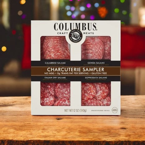The Columbus Charcuterie Sampler (12 oz) by Colombus elegantly rests on a wooden table, featuring Calabrese, Genoa, Italian Dry, and Sopressata salame. Blurred lights in the background add warmth to this exquisite cured meats collection.
