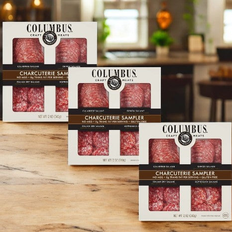 Three packages of Colombus Charcuterie Sampler (12 Oz- 3 Count) are elegantly displayed on a wooden table against a blurred kitchen background, each offering an exquisite variety of cured meats perfect for culinary enthusiasts.