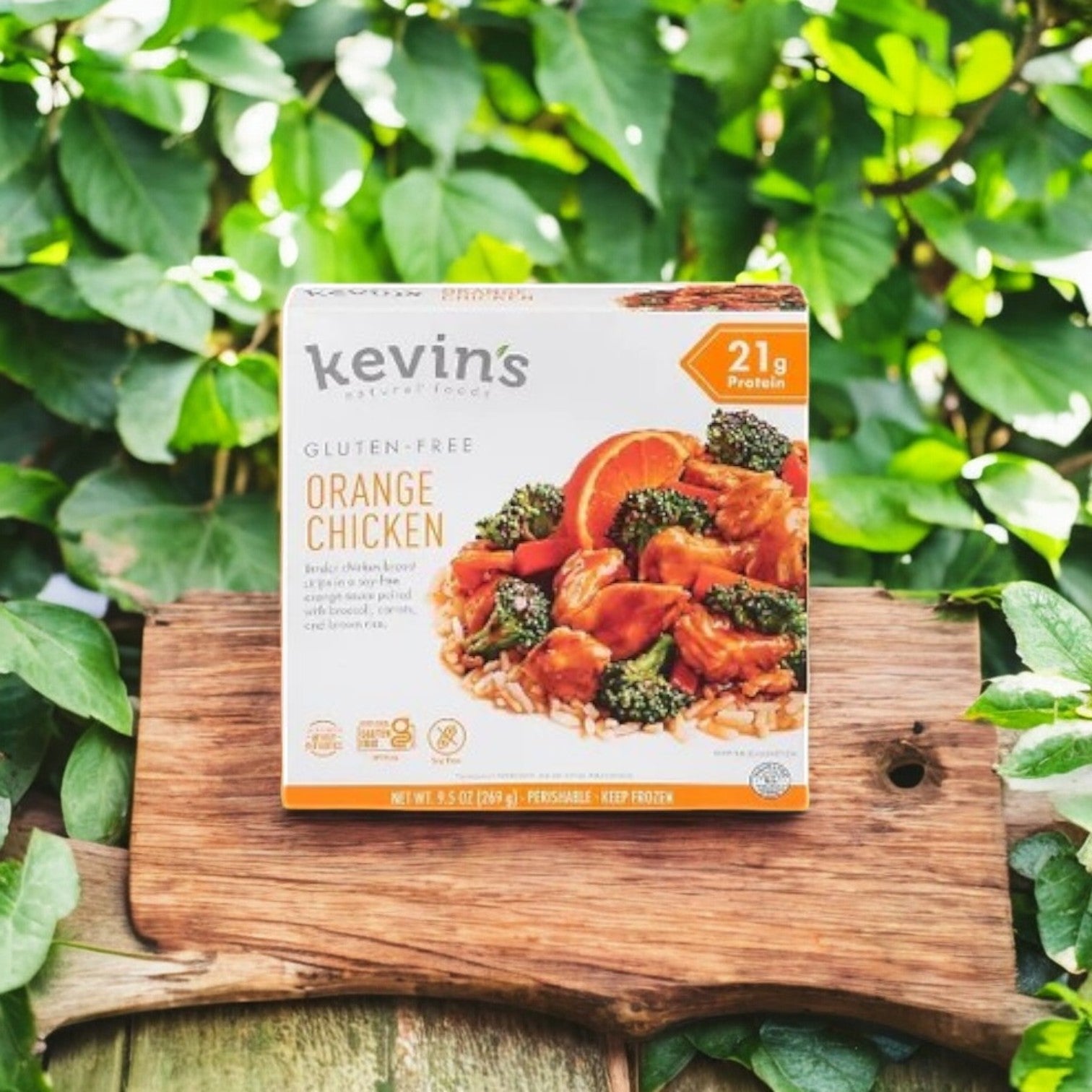 Kevin's 4 Pack Combo - Gluten-Free Gourmet Meals, including a 9.5 oz Orange Chicken, is showcased outdoors on a wooden board amid lush green leaves.
