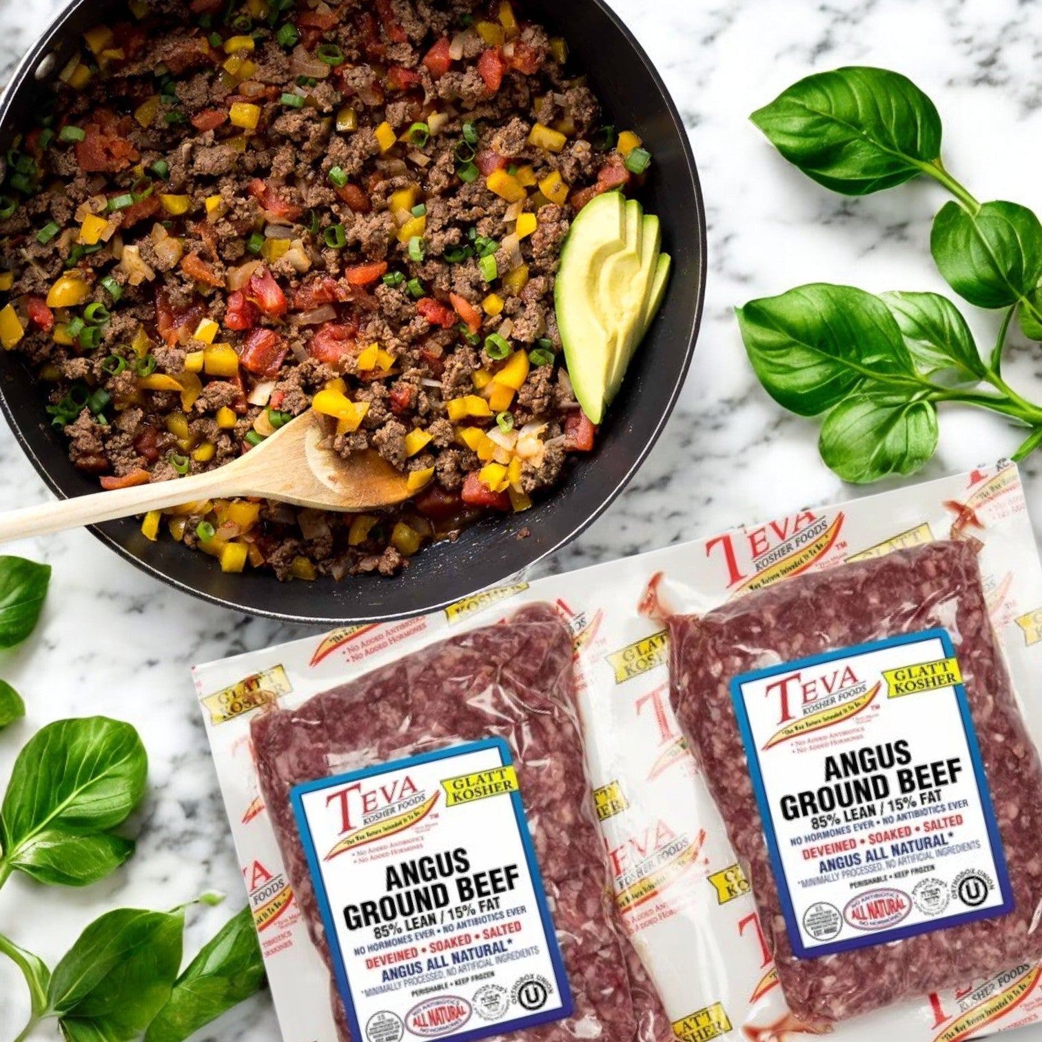 A skillet with cooked ground beef, peppers, and avocado slices sits beside two packages of Teva Kosher Foods Glatt Kosher Angus Ground Beef (85% lean) near basil leaves on a marble surface.