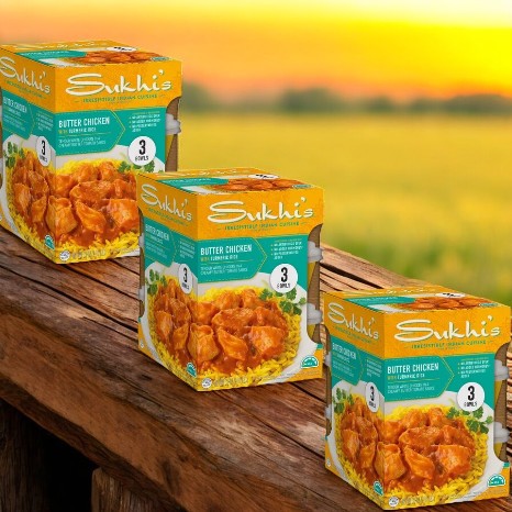 Three packs of Sukhi's Butter Chicken with Turmeric Rice rest on a wooden table, framed by a blurred field in the background.