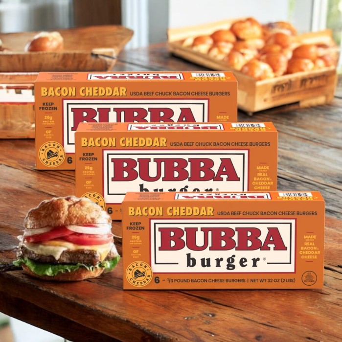 Three boxes of Bubba Burger Cheese Burgers, Bacon Cheddar- 3 Count, are on a wooden table with a bacon cheddar cheeseburger in the foreground on a cutting board, highlighting the savory appeal of these Bubba frozen patties.