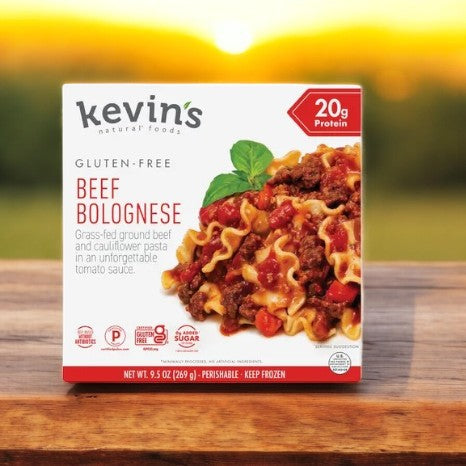 A box of Kevin's Natural Foods Beef Bolognese - 1 Count, showcasing an image of beef and pasta in tomato sauce. The packaging highlights its gluten-free label with 20g of protein and rests on a wooden surface against a blurred outdoor backdrop. Brand: Kevin's.
