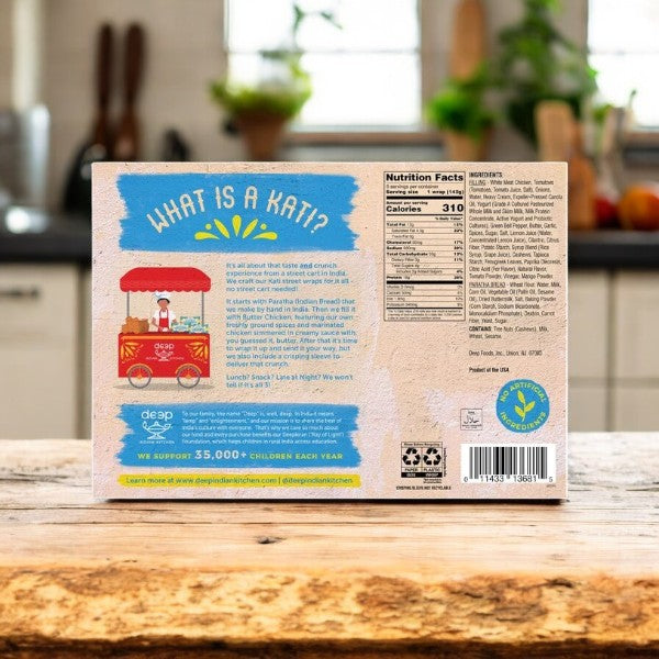 The back of a Deep Indian Kitchen Kati Street Wrap, Butter Chicken package sits on a wooden kitchen counter, displaying nutrition facts, βWhat is a Kati?β, colorful graphics, and an intro to this frozen meal inspired by Indian street food.