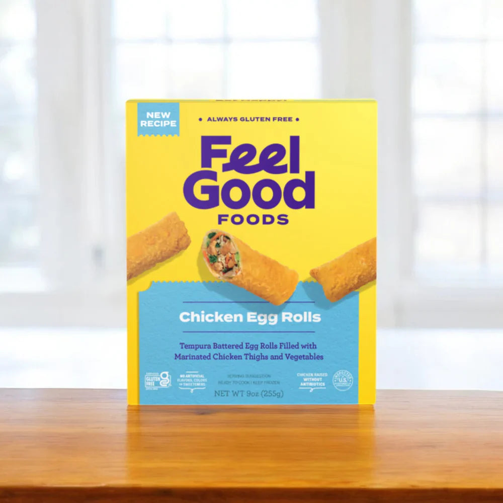 A vibrant yellow Feel Good Foods Gluten-Free Bundle box sits on a wooden surface, displaying "New Recipe" and "Always Gluten Free" labels. Whether enjoying crispy Chicken Egg Rolls or savory potstickers, every bite promises pure comfort.