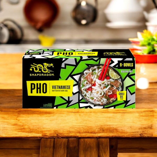 A 2.7 oz box of Snapdragon Vietnamese Pho, Beef flavor, from a 9-count pack is pictured with a bowl of noodle soup, beef slices, and vegetables on the packaging, displayed on a wooden kitchen table.