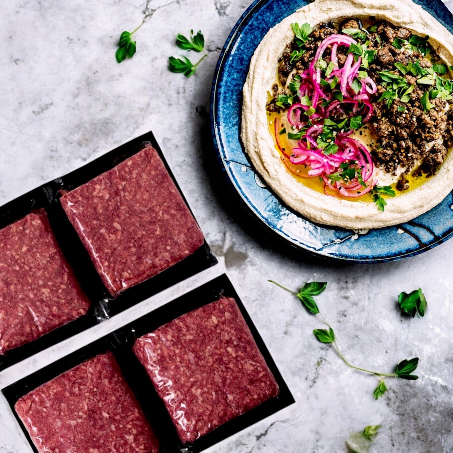 Four packs of Diamond Valley Halal Ground Beef, 85% Lean – 1 lb Chub (4 Pack) by New Zealand Spring Lamb sit beside a blue plate of hummus topped with seasoned meat, pickled onions, and herbs on a gray marble surface, all ready to complement your premium tuna seafood dishes.